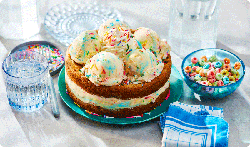 aqua cake plate with chocolate chip cookie birthday cake ice cream cake with scoops of ice cream and whipped cream on top, and a side bowl of fruit hoops cereal