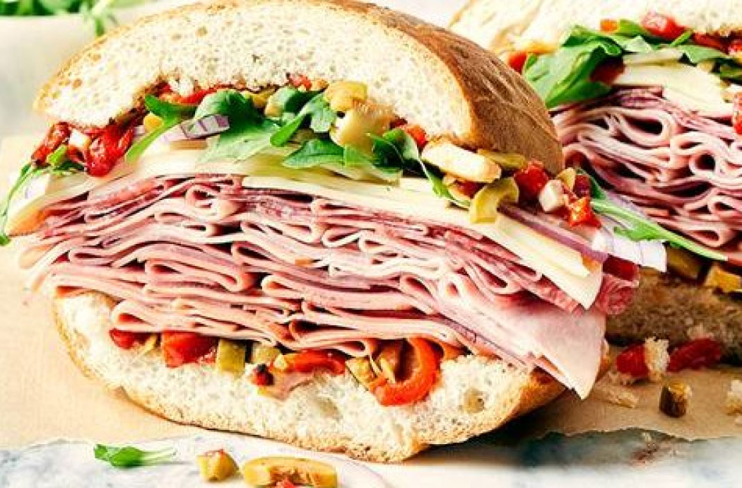 Deli meat, cheese, olive, roasted peppers, red onion, and arugula on a French loaf, sitting on a marble chopping block with next to a bowl of chopped, pickled vegetables.