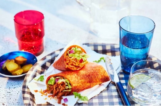 Red pepper wrap surrounding a maple bacon pulled pork filling with lettuce, red onions, cheddar and bbq sauce on a blue and white gingham plate next to a yellow napkin.