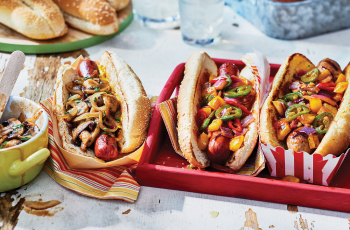 A table with a red platter of topping-laden hot dogs and napkins and side dishes of toppings and hot dog buns.