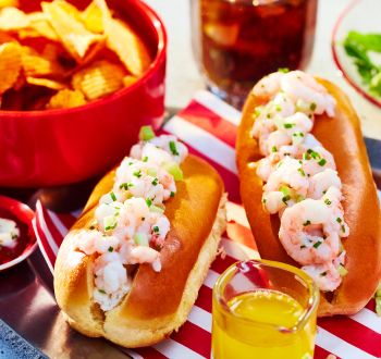 Two shrimp roll sandwiches in hot dog buns on a silver serving platter lined with a red and white sheet of parchment paper