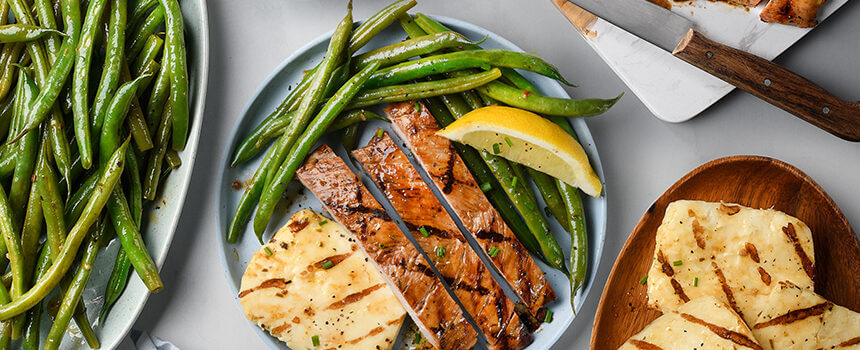 A table is filled with plates of Grilled Herb and Garlic pork tenderloin served with green beans, halloumi cheese and lemon wedge.