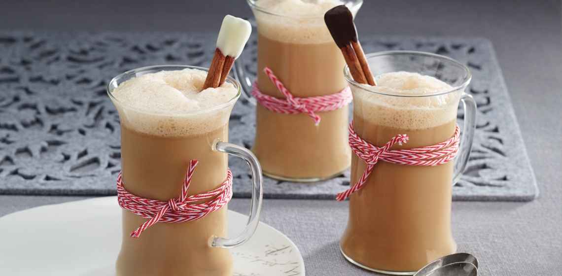 A grey table with three clear mugs of Gingerbread Almond Latte with cinnamon stir sticks and red and white twine wrapped around the middle.