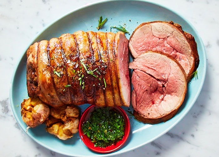 Top-down view of a light blue plate of boneless leg of lamb with green mint sauce on the side.