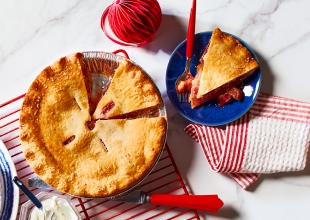 White marble surface with red cooling rack, holding one strawberry rhubarb pie with a slice cut out, and a blue side plate with pie slice and red fork