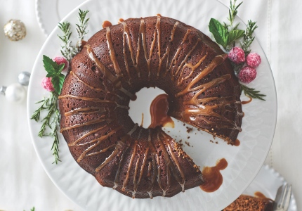 Overhead looking down at a white holiday tabletop with a white cake stand with a gingerbread Bundt cake drizzled with sauce, green and red garnish, and once slice cut out.