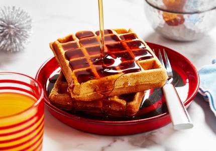 A close-up of waffles on a red plate with syrup being drizzled from up high, and a fork on the side.