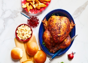 a top-do red plate with potato wedges and stuffing, a blue plate with a whole roasted chicken, and a side dish of macaroni salad, cranberry sauce, and fresh baked buns