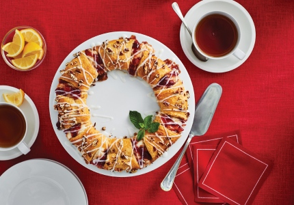 A strawberry crescent-roll wreath with an icing sugar glaze drizzle on a red serving platter with mugs of coffee and side plates.