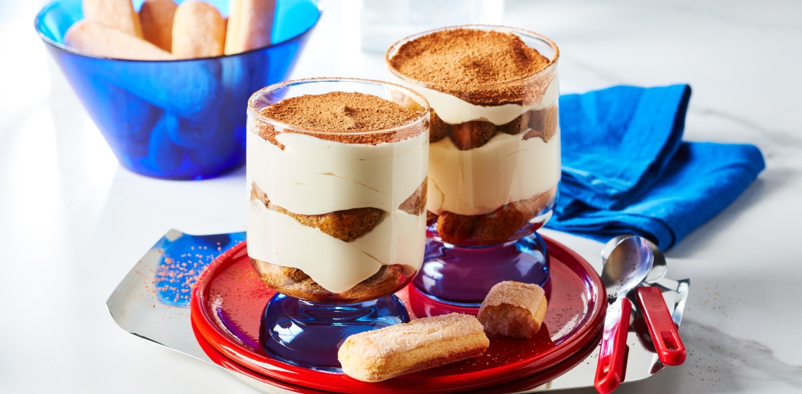 Small red serving tray with two Individual cups of tiramisu trifle, layered with espresso-soaked ladyfingers, mascarpone cream and cocoa sprinkle.
