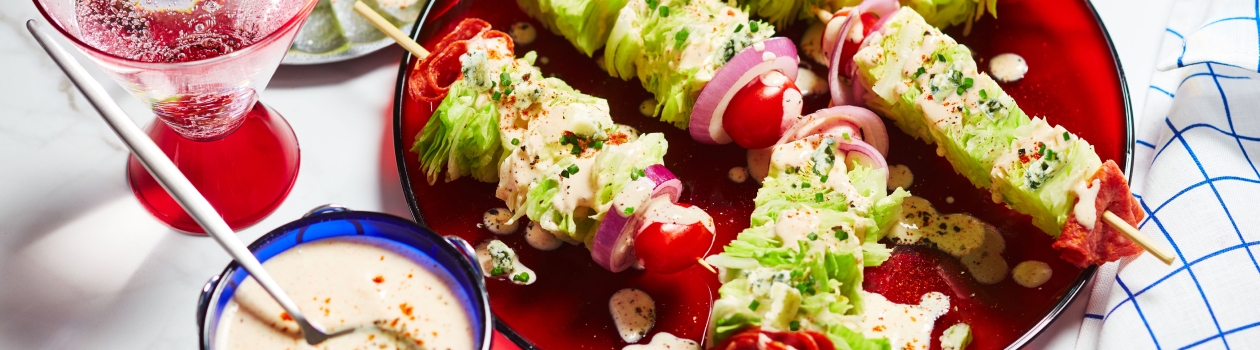 Red serving platter with five wedge salad skewers, and a side dish of dressing with a spoon dipped in