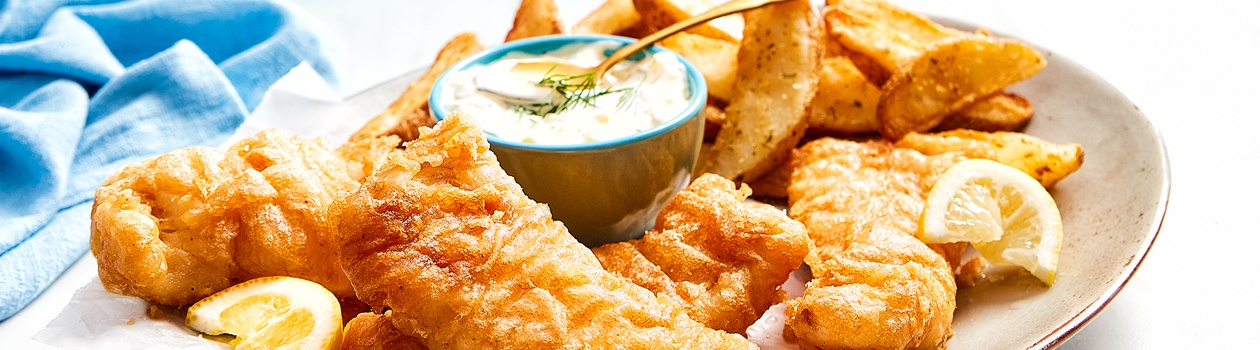 cream coloured oval platter with beer battered fried fish, French fries, lemon wedges and tartar sauce in a side bowl.