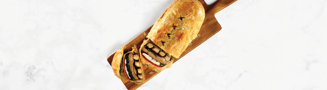 Wooden cutting board with partially sliced portobello mushroom wellington, with slices showing inside layers of spinach, mushroom and sausage.